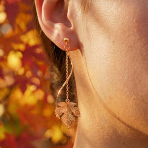 May include: A gold-toned dangle earring featuring a delicate chain and a spiraling metal wire element that terminates in a detailed, textured maple leaf charm. The earring is attached to a small ball stud post, creating a long, slender silhouette.