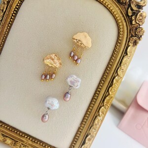 May include: A pair of gold cloud earrings with dangling pink pearls. The earrings are displayed on a gold-framed velvet board.