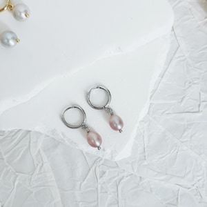 May include: A pair of silver hoop earrings with a single pink pearl dangling from each hoop.