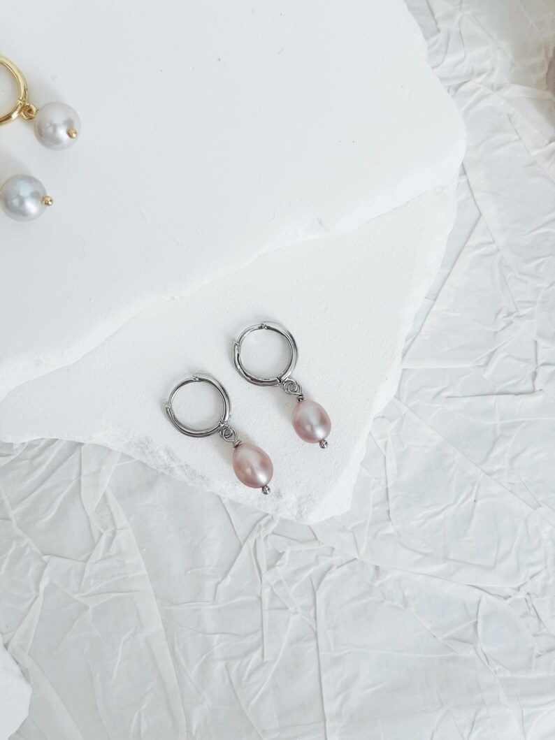 May include: A pair of silver hoop earrings with a single pink pearl dangling from each hoop.