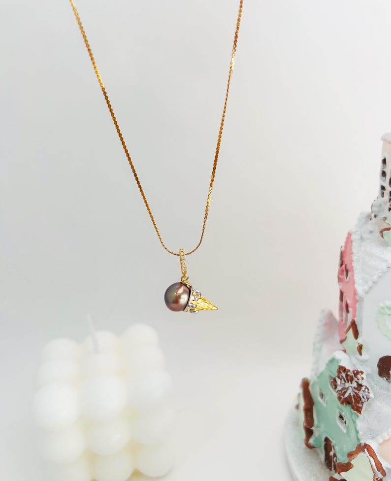 K&ouml;nnte beinhalten: Eine goldene Kettenkette mit einem Anh&auml;nger in Form eines goldenen Eisbechers mit einer dunkelbraunen Perle und kleinen klaren Steinen.