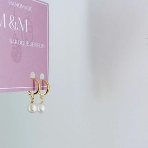 May include: A pair of gold hoop earrings with a single white pearl dangling from each hoop. The earrings are displayed on a pink card with the text "HANDMADE M&M BAROQUE JEWELRY".