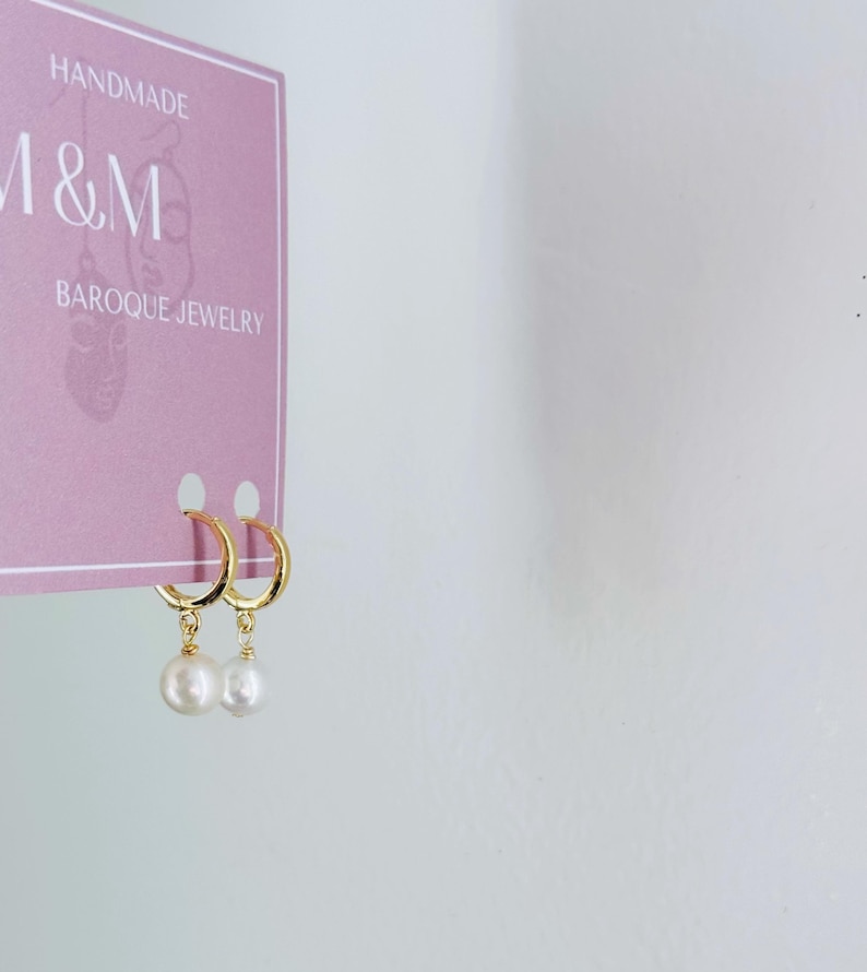 May include: A pair of gold hoop earrings with a single white pearl dangling from each hoop. The earrings are displayed on a pink card with the text "HANDMADE M&M BAROQUE JEWELRY".