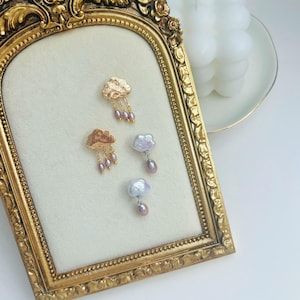 May include: A pair of gold cloud earrings with pink pearl drops and a pair of pearl cloud earrings with pink pearl drops displayed on a gold frame.