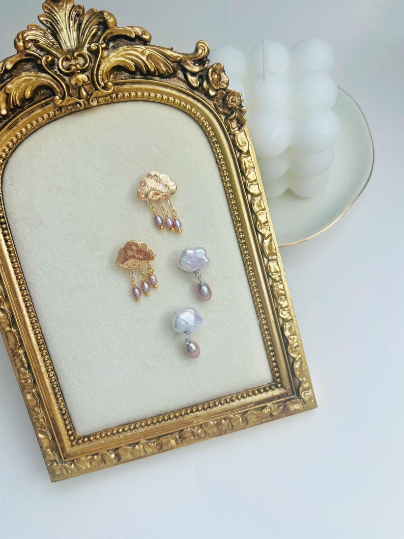 May include: A pair of gold cloud earrings with pink pearl drops and a pair of pearl cloud earrings with pink pearl drops displayed on a gold frame.