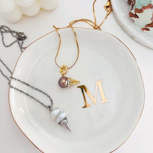 K&ouml;nnte beinhalten: Zwei goldene Halsketten mit Perlenanh&auml;ngern auf einer wei&szlig;en Keramik-Schale mit goldfarbenem Rand. Die Schale hat den Buchstaben "M" in Gold auf der Oberfl&auml;che. Ein Anh&auml;nger ist eine einzelne dunkle Perle mit einem goldenen Blattdesign. Der andere Anh&auml;nger ist eine doppelte Perle mit einem silbernen Kegeldesign.