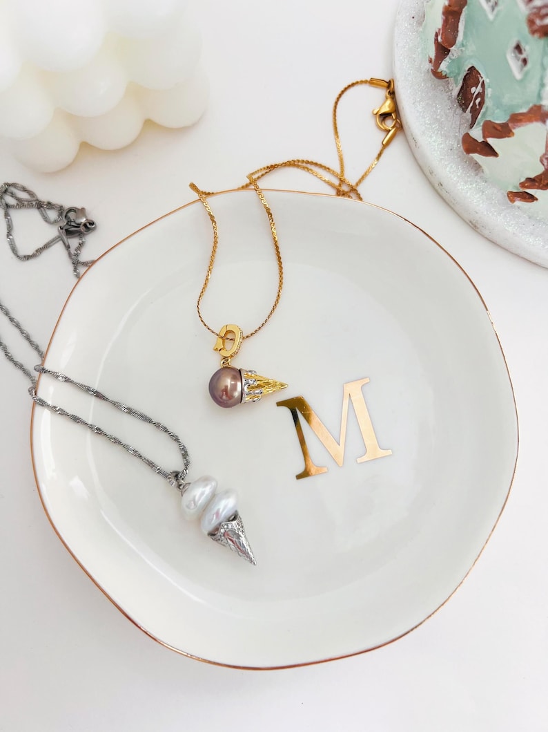 K&ouml;nnte beinhalten: Zwei goldene Halsketten mit Perlenanh&auml;ngern auf einer wei&szlig;en Keramik-Schale mit goldfarbenem Rand. Die Schale hat den Buchstaben "M" in Gold auf der Oberfl&auml;che. Ein Anh&auml;nger ist eine einzelne dunkle Perle mit einem goldenen Blattdesign. Der andere Anh&auml;nger ist eine doppelte Perle mit einem silbernen Kegeldesign.