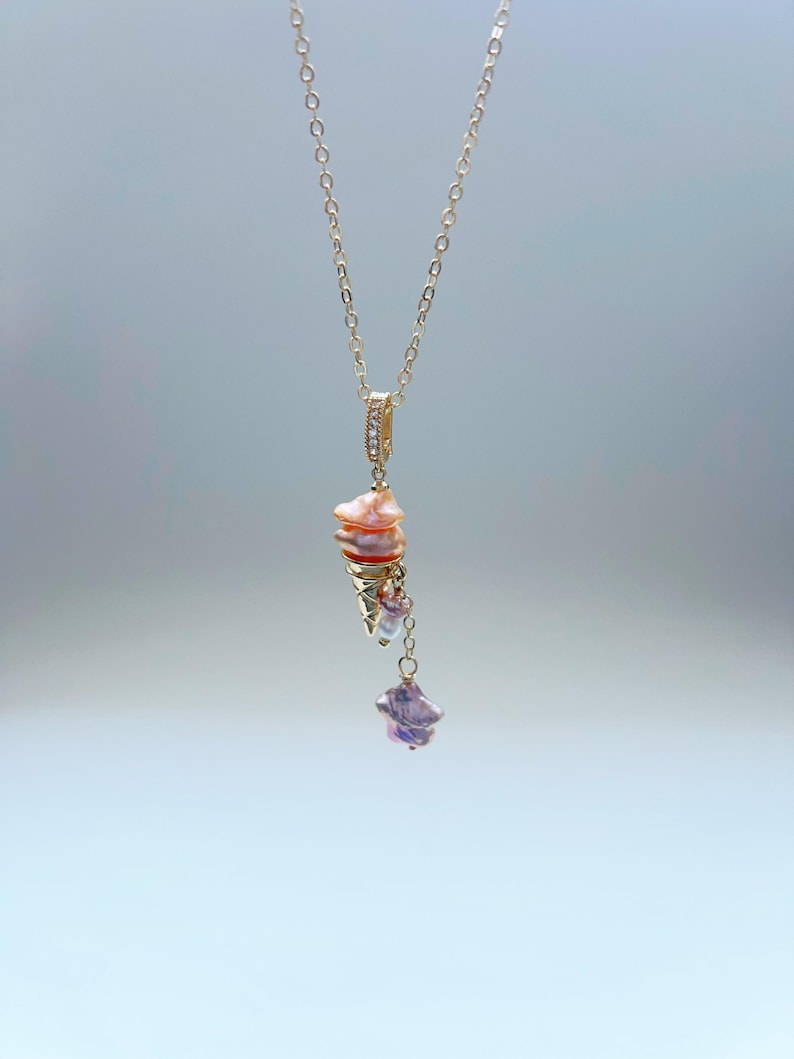 K&ouml;nnte beinhalten: Eine zarte goldene Kettenkette mit einem Anh&auml;nger aus rosa und wei&szlig;en Perlen. Der Anh&auml;nger hat eine sternf&ouml;rmige Perle mit einer kleineren Perle, die daran h&auml;ngt.