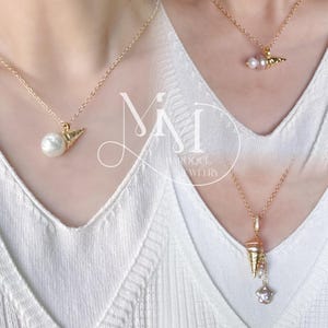 K&ouml;nnte beinhalten: Zwei goldene Halsketten mit Perlen- und Muschel-Anh&auml;ngern. Die Halskette links hat eine gro&szlig;e Perle und einen goldenen Muschel-Anh&auml;nger. Die Halskette rechts hat eine kleinere Perle, eine rosafarbene Perle und einen goldenen Muschel-Anh&auml;nger mit einer kleineren Perle, die daran h&auml;ngt.