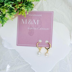May include: Gold hoop earrings with a single pearl dangling from each hoop. The earrings are displayed on a pink card with the text "HANDMADE M&M BAROQUE JEWELRY".