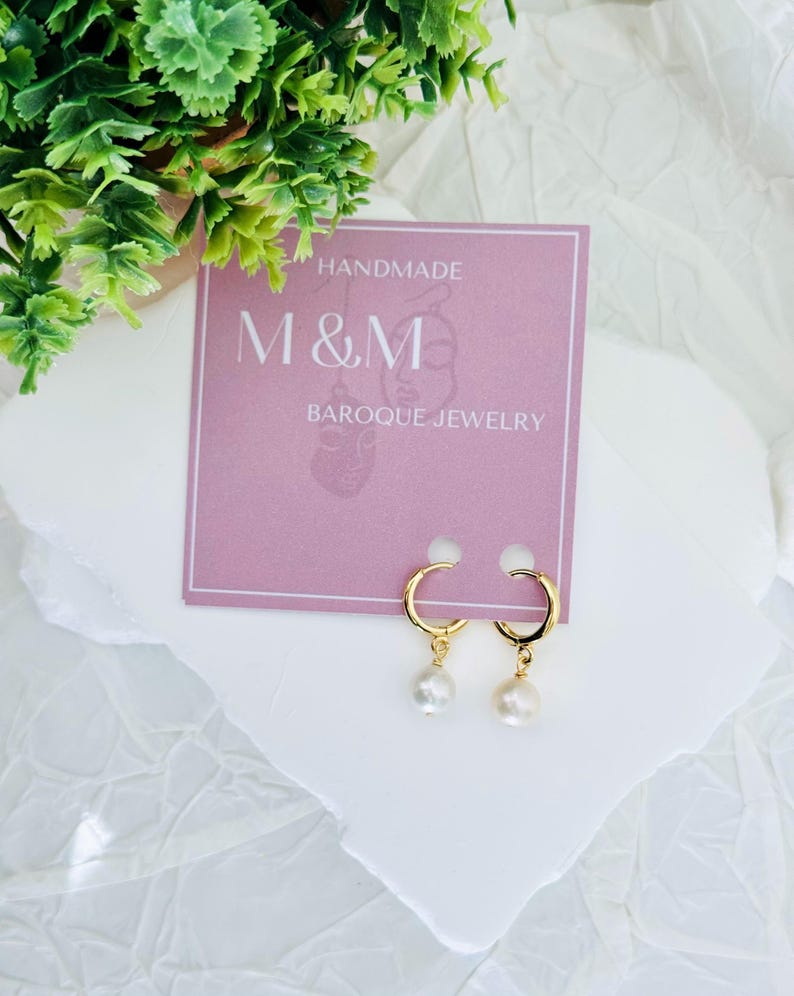 May include: Gold hoop earrings with a single pearl dangling from each hoop. The earrings are displayed on a pink card with the text "HANDMADE M&M BAROQUE JEWELRY".