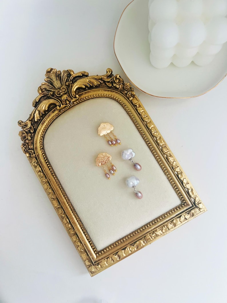 May include: Gold cloud earrings with pearl drops. The earrings are displayed on a gold ornate frame.