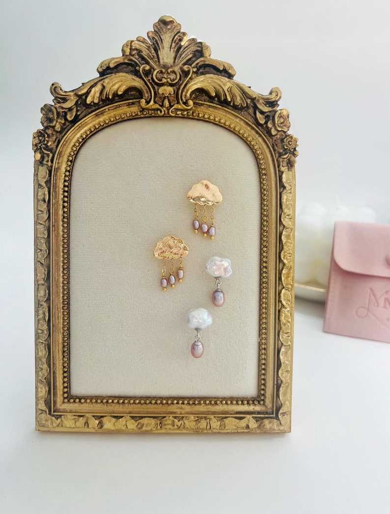 May include: A pair of gold cloud earrings with dangling pearls, displayed on a gold-framed stand. The earrings feature a delicate cloud design with a cluster of pearls hanging from each cloud.