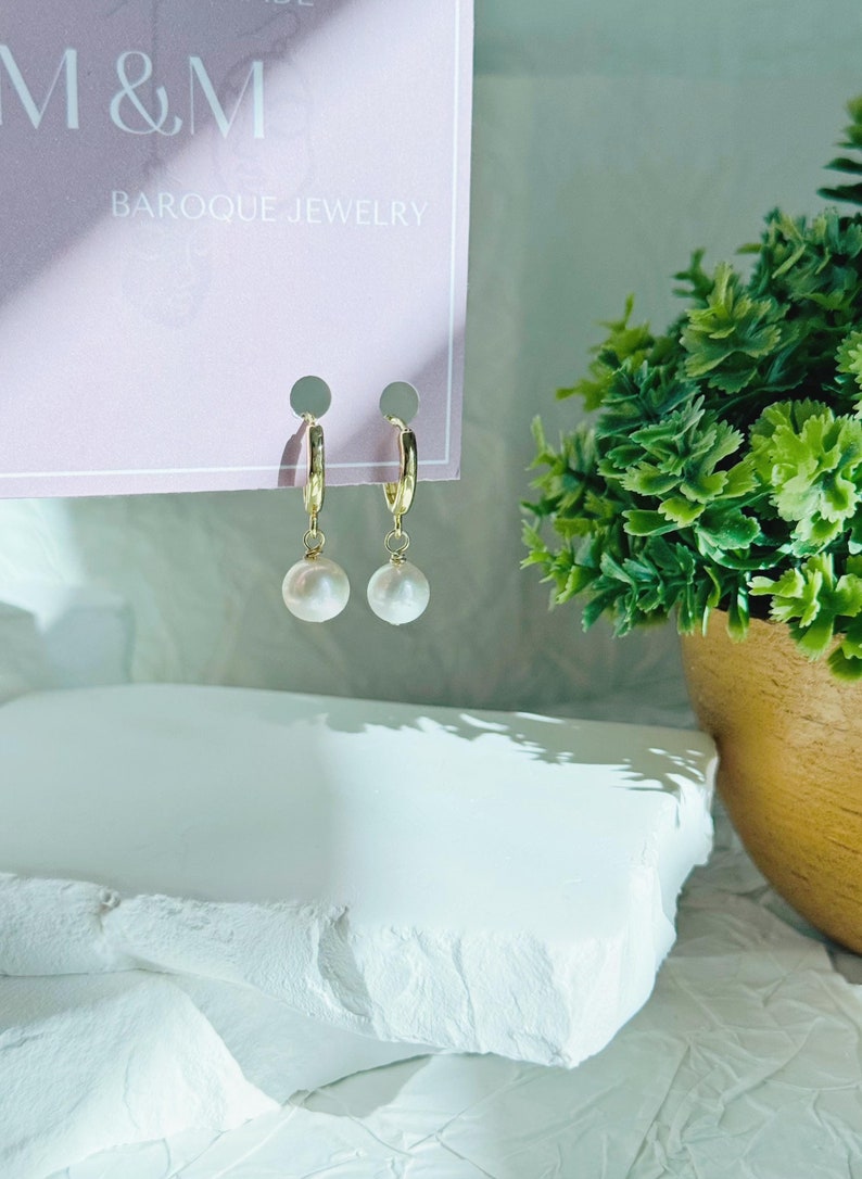 May include: A pair of gold hoop earrings with a white pearl dangling from each hoop. The earrings are displayed on a pink card with the text "M&M Baroque Jewelry".