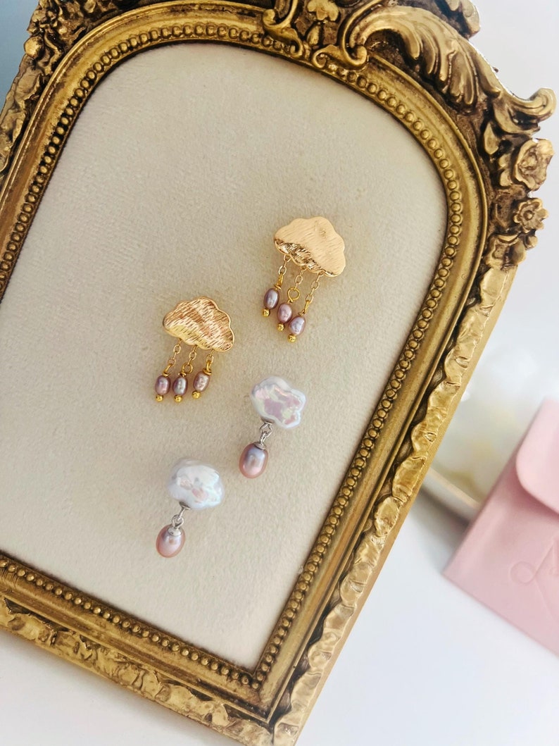 May include: A pair of gold cloud earrings with dangling pink pearls. The earrings are displayed on a gold-framed velvet board.