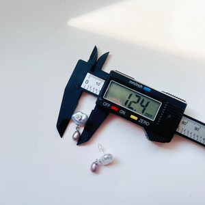 May include: A pair of silver earrings with a large, irregular shaped pearl and a smaller, round pearl hanging from it. The earrings are being measured with a digital caliper, which reads 12.4 mm.
