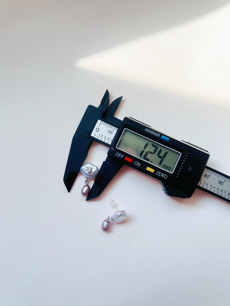 May include: A pair of silver earrings with a large, irregular shaped pearl and a smaller, round pearl hanging from it. The earrings are being measured with a digital caliper, which reads 12.4 mm.