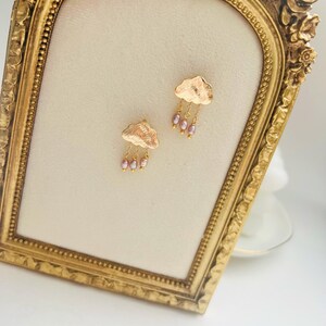 May include: Gold cloud earrings with pink pearl accents hanging from the bottom of each cloud.