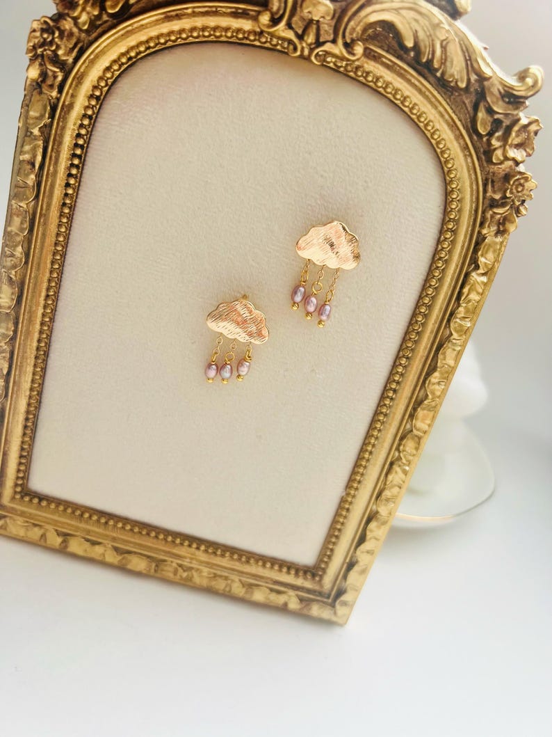 May include: Gold cloud earrings with pink pearl accents hanging from the bottom of each cloud.