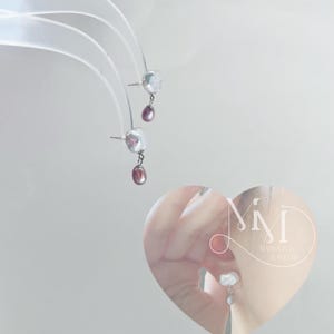 May include: A pair of silver earrings with a white pearl and a pink pearl dangling from each. The earrings are shown on a white background and in a heart-shaped frame with the text "Mimi Baroque Jewelry".