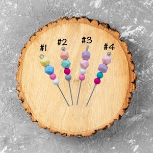 Peut inclure: Quatre brochettes décoratives avec des perles colorées sont présentées sur une surface en bois. Chaque brochette présente une combinaison différente de perles géométriques et rondes aux couleurs pastel, comme le rose, le bleu, le jaune et le turquoise. Les brochettes sont numérotées de 1 à 4.