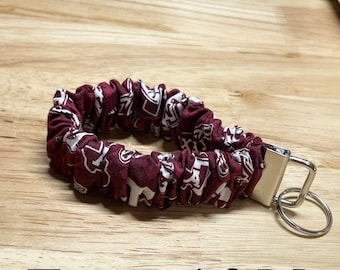 May include: Maroon and white patterned fabric wristlet keychain with a silver-colored metal clasp and key ring. The fabric is gathered and looped into a circular shape. The keychain rests on a light brown wooden surface.