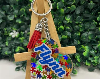 May include: A heart-shaped keychain with a red tassel and a silver chain. The heart is filled with colorful puzzle pieces and the name "Flynn" is written in blue letters.