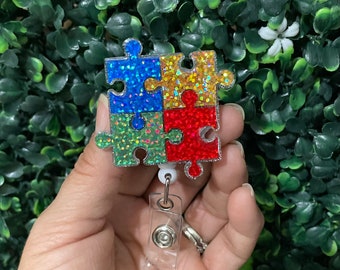 May include: A colorful glitter puzzle piece badge reel. The puzzle pieces are blue, green, yellow, and red. The reel is clear plastic with a silver metal clip.