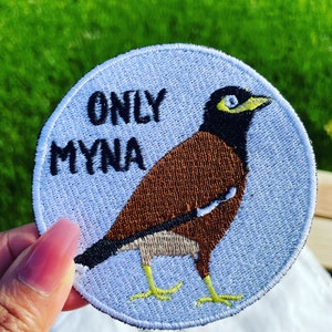 Puede incluir: Un parche bordado azul y blanco con un pájaro marrón y negro. El pájaro tiene un pico amarillo y un ojo amarillo. El texto "ONLY MYNA" está bordado en negro en el parche.