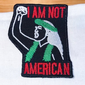 May include: Black and green embroidered patch with a white outline of a person with their fist raised in the air. The text "I AM NOT AMERICAN" is in red letters.