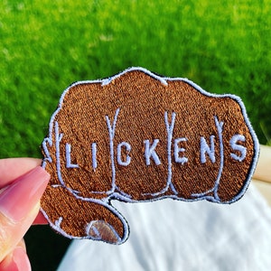 Könnte beinhalten: Ein brauner gestickter Aufnäher in Form einer Faust mit dem Wort "LICKENS" auf den Knöcheln.