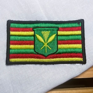 May include: An embroidered patch featuring the Hawaiian flag. The flag is green, yellow, and red with a green shield in the center. The shield has a yellow symbol of a spear and a feather.