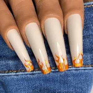 May include: Five long, almond-shaped, white acrylic nails with orange flame designs at the tips.