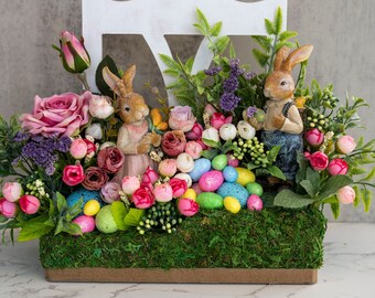 May include: A white wooden chair with a floral arrangement featuring pink and purple flowers, green leaves, and two bunny figurines. The arrangement is decorated with pastel-colored Easter eggs.