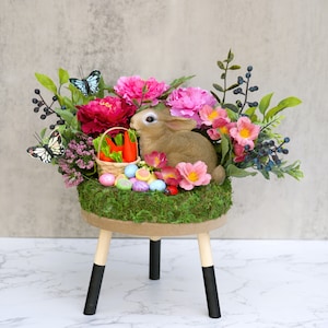 May include: A small wooden stool with black legs is decorated with a floral arrangement featuring pink and white flowers, green leaves, a brown bunny, and a basket of carrots. The arrangement is surrounded by colorful Easter eggs.