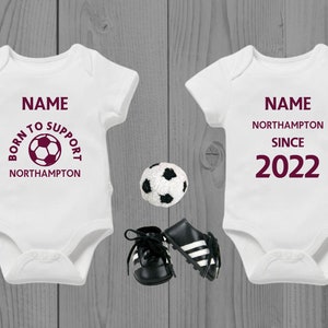 May include: Two white baby onesies with maroon text. One onesie says "Born to Support Northampton" with a football graphic. The other onesie says "Name Northampton Since 2022".