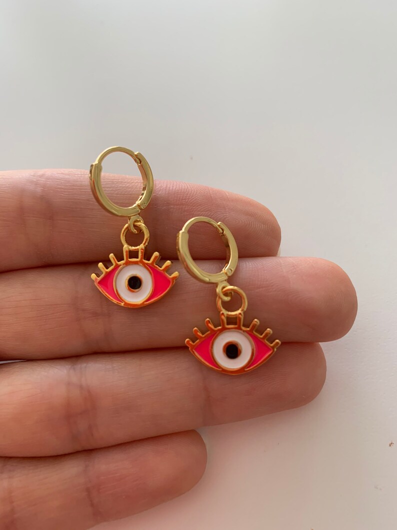 May include: A pair of gold hoop earrings with pink and white evil eye charms.