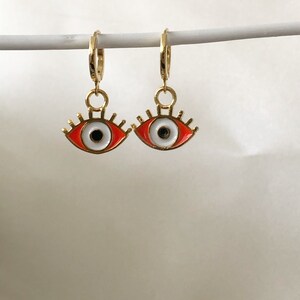 May include: A pair of gold hoop earrings with red and white evil eye charms. The charms have a white iris and black pupil with gold eyelashes.