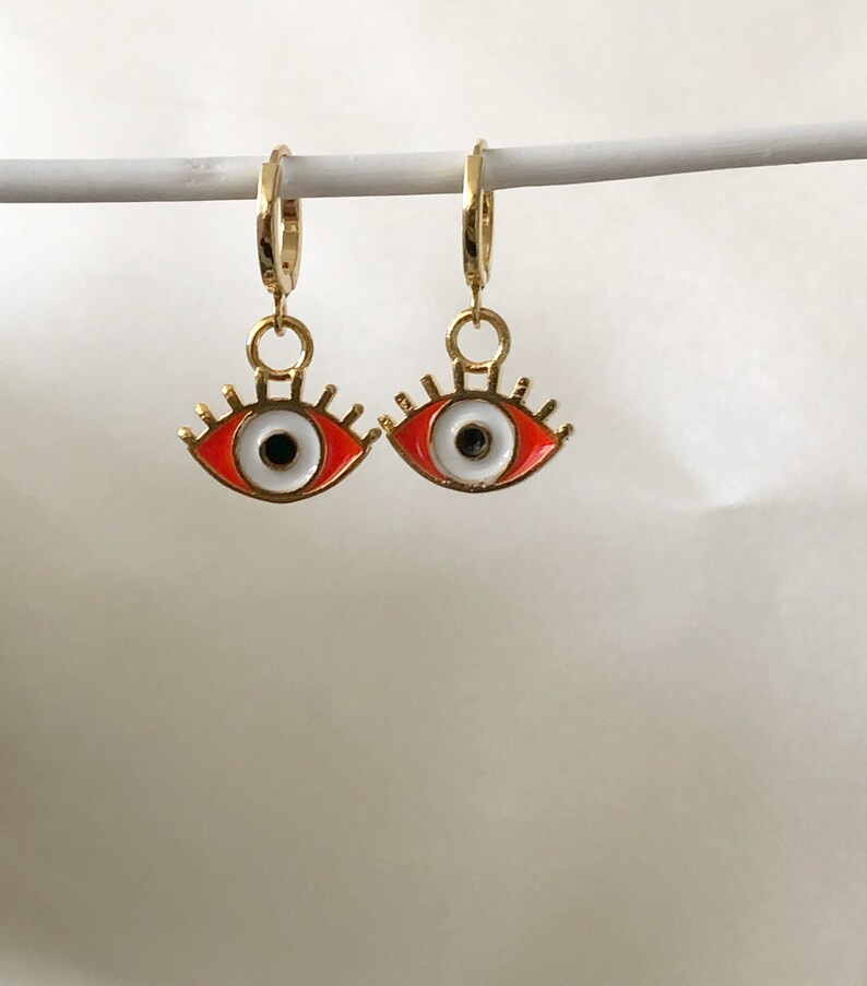 May include: A pair of gold hoop earrings with red and white evil eye charms. The charms have a white iris and black pupil with gold eyelashes.