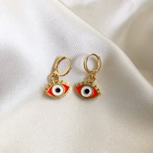May include: A pair of gold hoop earrings with red and white evil eye charms. The earrings are on a white background.