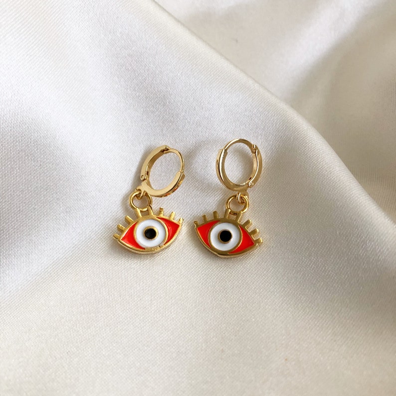 May include: A pair of gold hoop earrings with red and white evil eye charms. The earrings are on a white background.