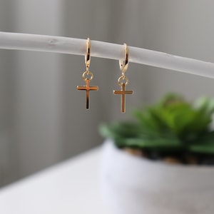 Peut inclure: Une paire de boucles d'oreilles créoles dorées avec un petit pendentif en forme de croix dorée suspendu à chaque créole.