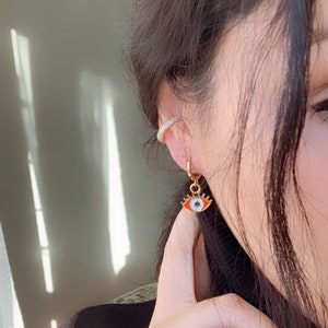 May include: Gold hoop earrings with a dangling red and white evil eye charm. The charm has a white pupil and a gold outline.