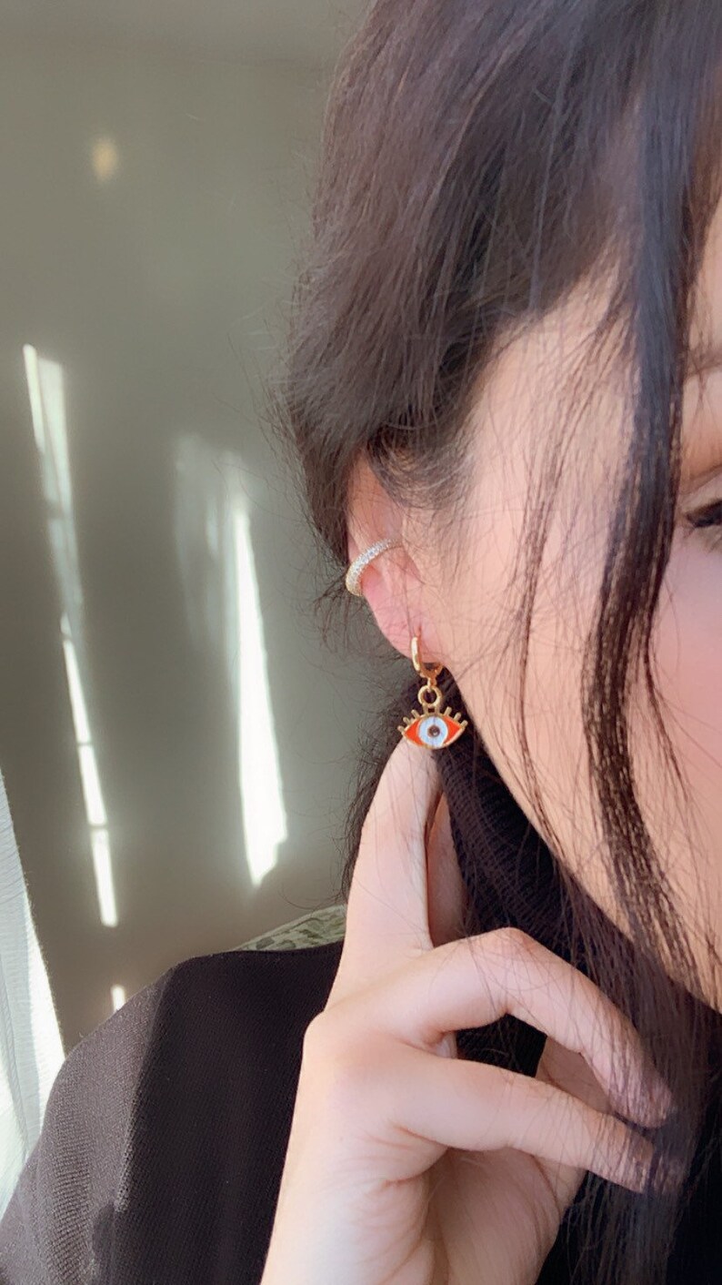 May include: Gold hoop earrings with a dangling red and white evil eye charm. The charm has a white pupil and a gold outline.