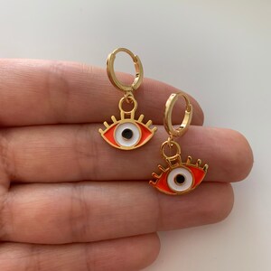 May include: A pair of gold hoop earrings with a dangling evil eye charm. The evil eye is red with a white iris and black pupil.