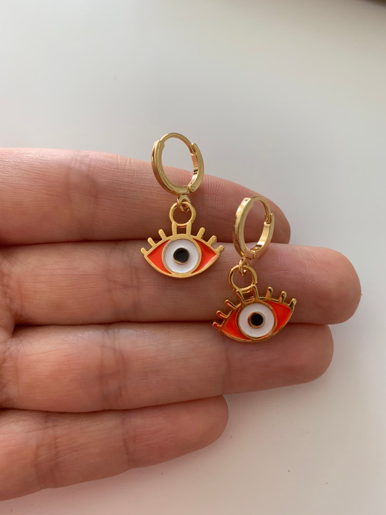 May include: A pair of gold hoop earrings with a dangling evil eye charm. The evil eye is red with a white iris and black pupil.