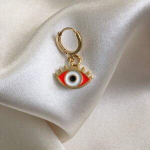 May include: Gold hoop earring with a red and white evil eye charm. The eye is outlined in gold and has a black pupil.