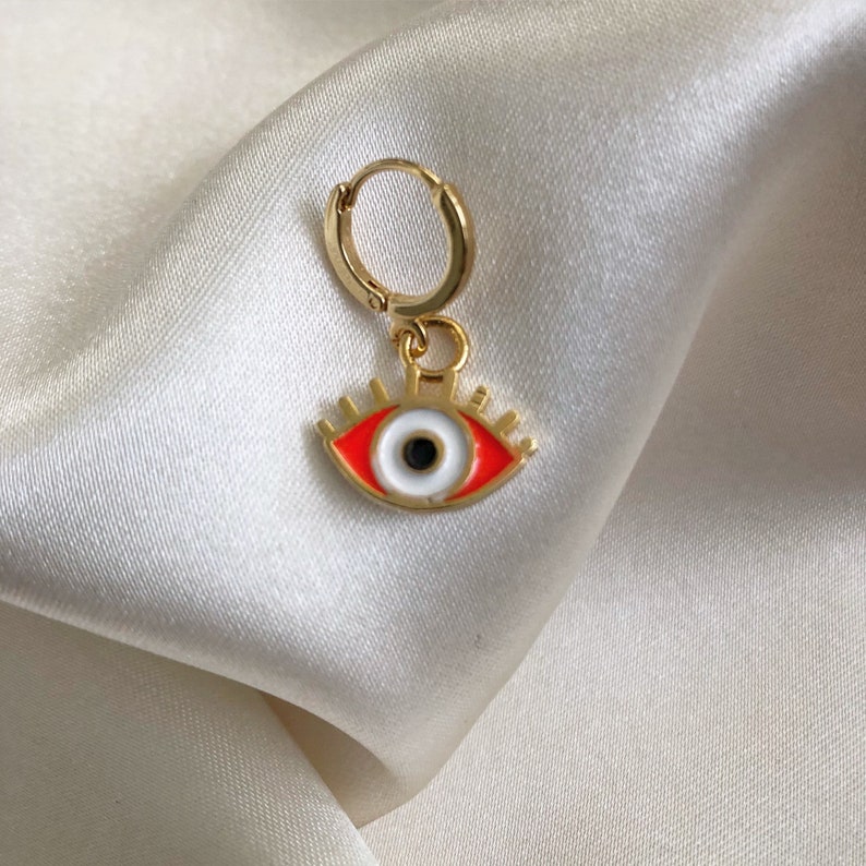 May include: Gold hoop earring with a red and white evil eye charm. The eye is outlined in gold and has a black pupil.