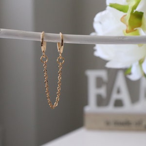 Peut inclure: Boucles d'oreilles créoles dorées reliées par une fine chaîne. Les boucles d'oreilles sont suspendues à une branche blanche.