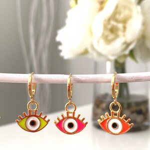 May include: Three gold hoop earrings with a dangling evil eye charm. The eyes are white with black pupils and have different colored rims: green, pink, and orange.